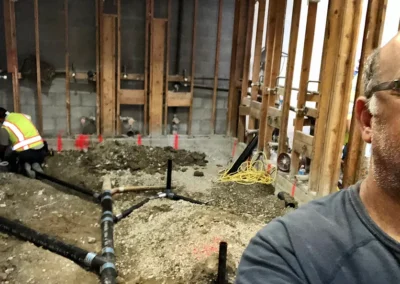 A man takes a selfie in a construction site, with a worker in a safety vest digging in a gravel area with exposed pipes and wooden framing.