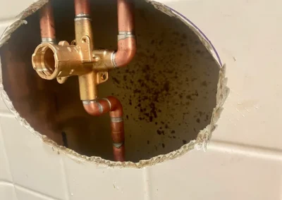 Close-up of a large hole in a wall revealing copper plumbing pipes and a brass fitting, surrounded by dark stains.