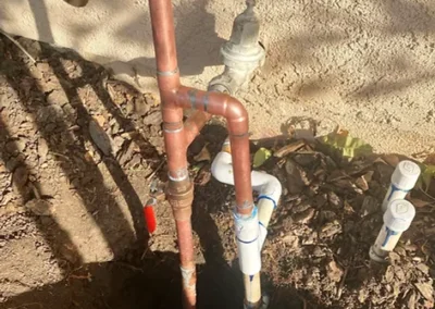 Copper and PVC pipes arranged around a dirt hole, featuring a faucet at the top and a water meter to the side.