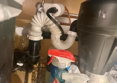 Under-sink plumbing featuring flexible pipes, a garbage disposal unit, and cleaning supplies, showcasing a typical kitchen setup.