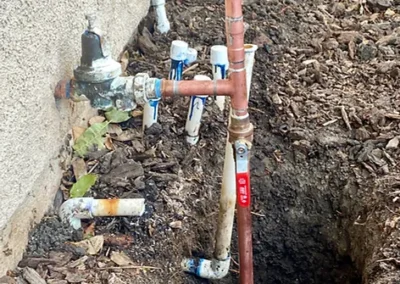 A cluster of pipes, including copper and PVC, is visible in a dirt trench next to a building’s foundation.