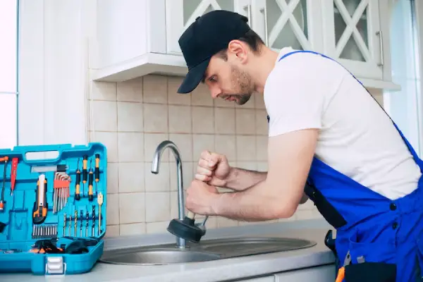 A maintenance worker in a blue uniform uses a plunger at a kitchen sink, with a tool kit filled with various tools beside him.